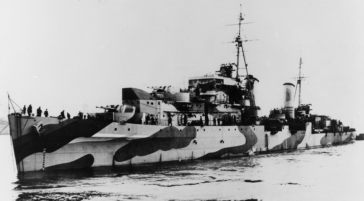 Black-and-white photograph of HMS Charybdis from the port bow, showing sailors on deck, forward guns, bridge, mast and camouflage.