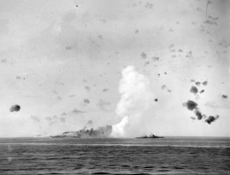 HMS Charybdis passes across the foreground, with the aircraft carrier Indomitable and a plume of smoke behind.