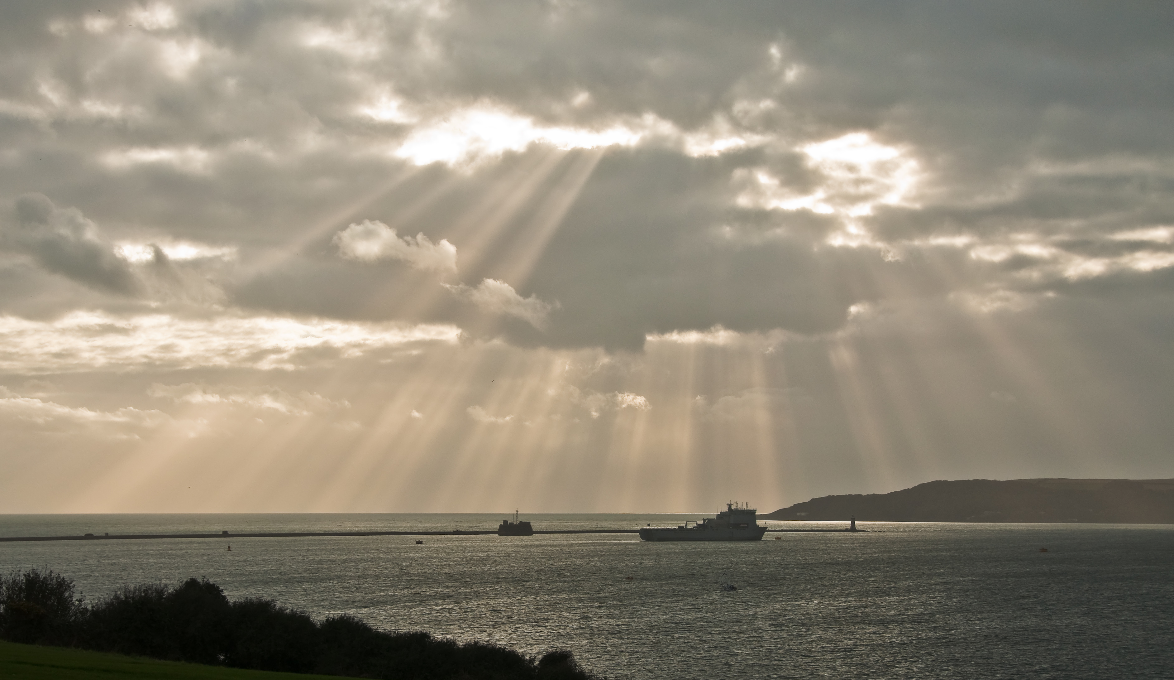 Sunbeams fall through a cloudy sky onto Plymouth Sound, its breakwater and a modern ship.