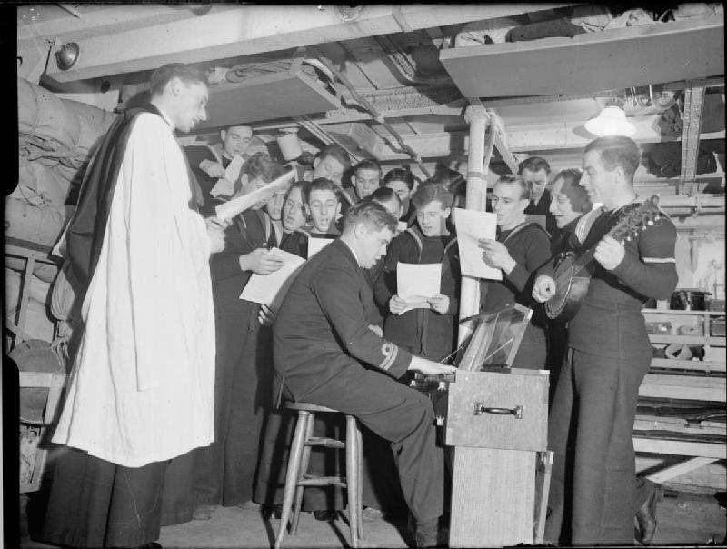Sailors gathered below deck around a harmonium, singing from printed sheets as a naval chaplain conducts.